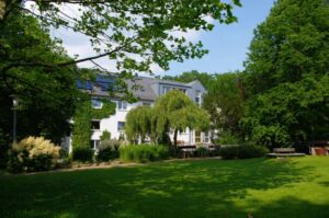 Helles Gebäude mit mehreren Fenstern, umgeben von einer gepflegten Grünanlage mit Bäumen und Rasen.