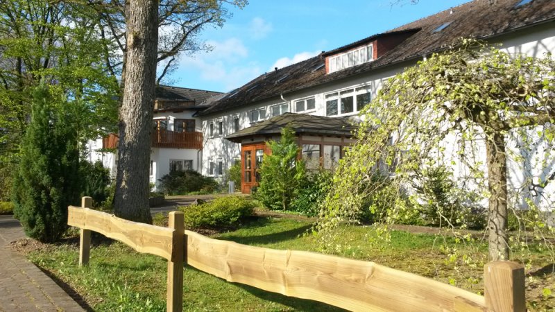 Helles Gebäude mit Holzeingang, umgeben von Gartenflächen, Bäumen und einem Holzzaun im Vordergrund.