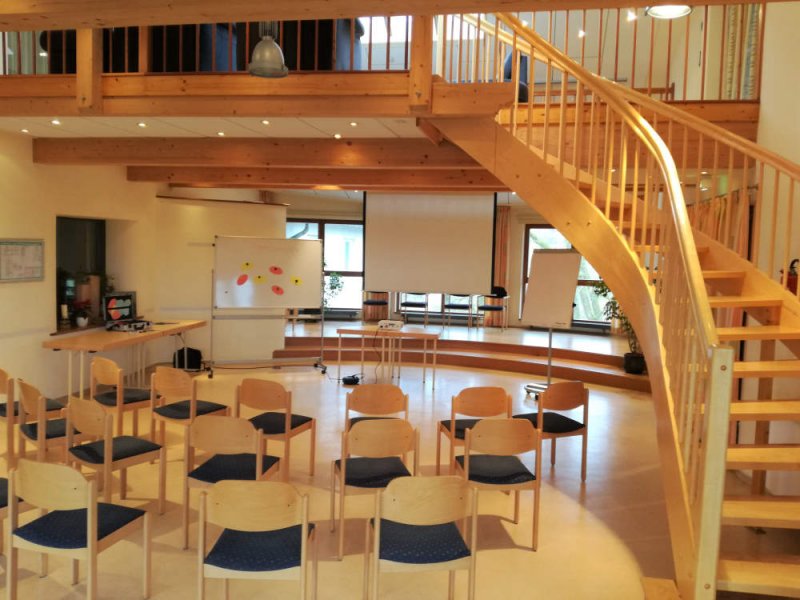 Veranstaltungsraum mit Treppe und Bestuhlung Großer Raum mit Holztreppe, Stuhlreihen und Präsentationsbereich.