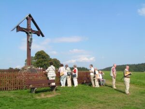 Mehrere Erwachsene stehen im Freien an einem hölzernen Wegkreuz auf einer Wiese.