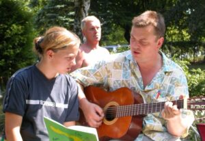 Erwachsene Person spielt im Freien Gitarre, daneben hält ein Kind ein aufgeschlagenes Buch.