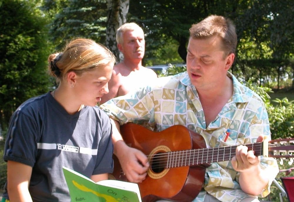 Erwachsene Person spielt im Freien Gitarre, daneben hält ein Kind ein aufgeschlagenes Buch.