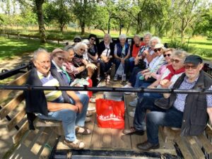 Mehrere ältere Erwachsene sitzen gemeinsam auf Sitzbänken eines offenen Anhängers im Grünen.