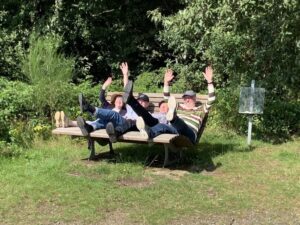 Mehrere Erwachsene sitzen oder liegen auf einer Bank im Grünen und heben die Arme.