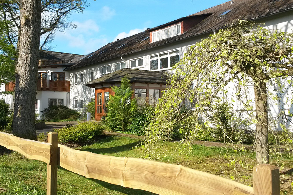 Weißes Gebäude mit mehreren Fenstern und Holzeingang, umgeben von Garten und Bäumen.