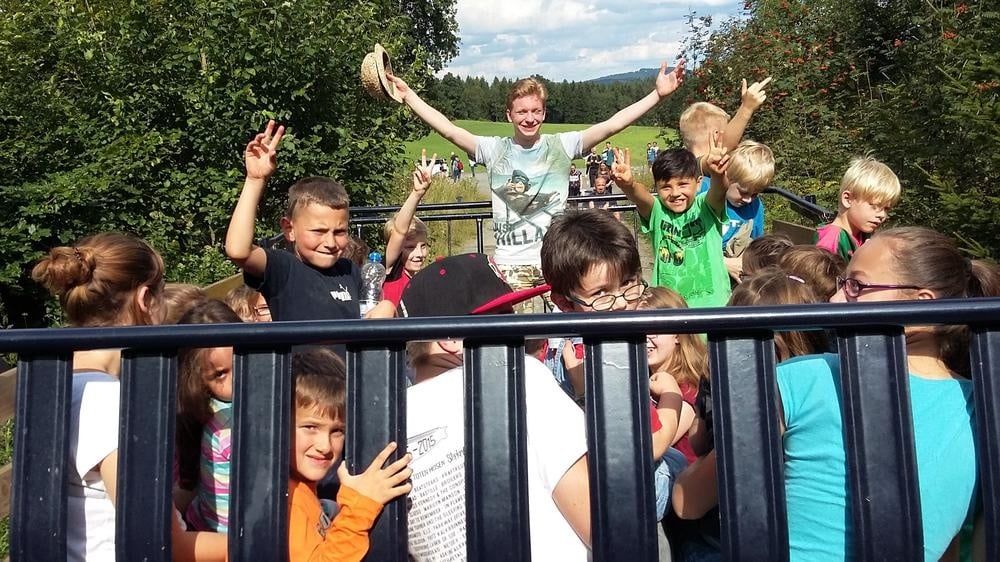 Gemeinsamer Ausflug mit Kindergruppe Gruppe von Kindern und einer betreuenden Person auf einem offenen Anhänger im Freien, mehrere Kinder heben die Hände.